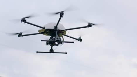 Handheld-camera-shot-of-drone-with-six-rotors,-white-body,-green-circuit-board,-camera,-and-two-black-landing-gears-in-flight-against-a-cloudy-sky