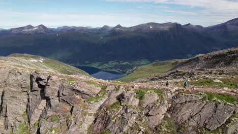 Dos-Excursionistas-Caminando-Al-Borde-De-Una-Impresionante-Cresta-En-Noruega,-Romdalseggen,-Vista-Aérea-De-Un-Paisaje-Montañoso-Con-Un-Fiordo-Al-Fondo
