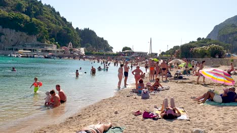 Gente-En-La-Playa-Nadando-Y-Tomando-El-Sol,-Clima-Ventoso-En-Palaiokastritsa-Corfu-Grecia,-Imágenes-En-Tiempo-Real