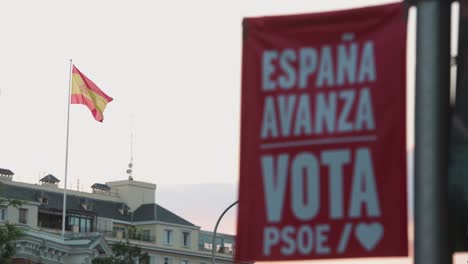 Ein-Banner-Der-Spanischen-Sozialistischen-Arbeiterpartei-Erinnert-Die-Öffentlichkeit-Daran,-Während-Der-Spanischen-Parlamentswahlen-Zu-Wählen,-Während-Im-Hintergrund-Eine-Spanische-Flagge-Weht