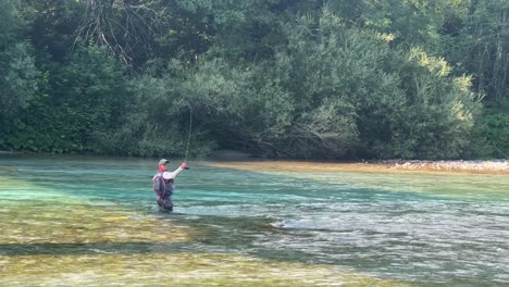 Man-is-fly-fishing-trout-on-Soca-river-on-sunny-day-in-Slovenia,-static-shot