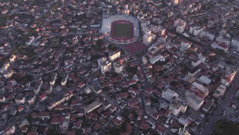 Reveal-shot-of-city-Shkodër-with-soccer-stadium-in-the-middle-with-sunrise,-aerial