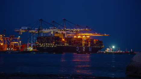 Timelapse-shot-of-large-blue-crane-loading-containers-over-a-large-container-ship-in-Kalafrana,-Birzebbuga,-Malta-from-night-to-early-morning-time