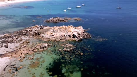 Aerial-Orbital-Footage-of-Rocky-Shore-with-Clear-and-Dark-Water-and-Sailboats