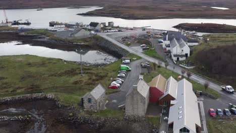 Tilting-drone-shot-of-the-Taigh-Chearsabhagh-Museum-and-Arts-Centre,-the-marina-and-the-mountains,-moor-and-peatland-beyond-the-bay-in-Lochmaddy