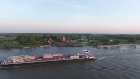 Container-vessel-on-river-waal-passing-by-Slot-Loevestein-castle,-aerial