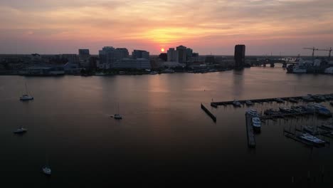 Flying-a-drone-along-a-marina-pier-with-the-boats-in-the-foreground-and-the-Norfolk-Virginia-Waterfront-in-the-background-with-the-rising-sun-peaking-out-from-behind-the-buildings-as-it-moves