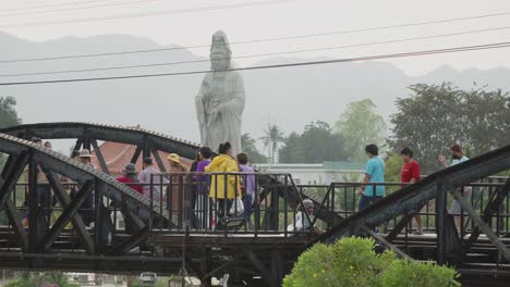 Tourists-walking-along-the-railway-tracks-on-the-Kwai-River-bridge-with-the-Buddhist-Guan-Yin-statue-in-the-background,-Kanchanaburi,-Thailand