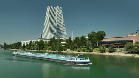 Oil-tanker-steaming-up-Rhine-river-Basel-with-roche-tower-and-Tinguely-museum-in-background
