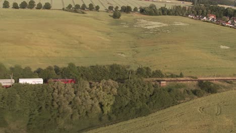 Drone-shot-of-a-red-and-white-train-hauling-white-cargo-containers-through-a-vast-green-field-interspersed-with-brown-patches,-with-houses-and-buildings-in-the-background-under-a-clear-blue-sky