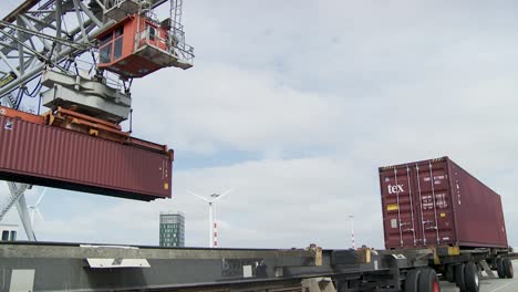 Ein-Großer-Roter-„Tex“-Transportcontainer-Wird-Von-Einem-Kran-Mit-Weiß-blau-Gestreiftem-Arm-Vor-Dem-Hintergrund-Eines-Wolkenverhangenen-Blauen-Himmels,-Eines-Gebäudes-Und-Von-Windkraftanlagen-Angehoben