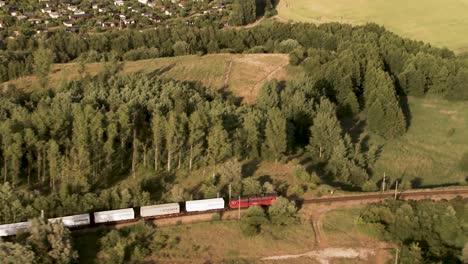 Drone-shot-reveals-a-red-and-white-cargo-train-traversing-a-lush-forest-area,-parallel-to-a-dirt-road-with-houses-and-rolling-fields-in-the-backdrop