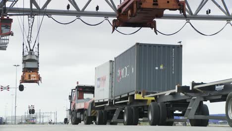 A-large-orange-"Stinis"-crane-lifts-a-gray-"Hapag-Lloyd"-shipping-container-off-a-red-"Hillebrand"-truck-at-a-bustling-port-under-a-cloudy-sky