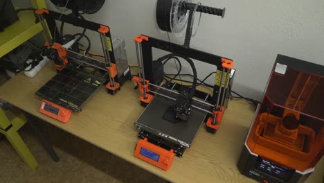 Three-3D-printers-on-the-table,-one-of-the-working-on,-orange-details,-black-filaments