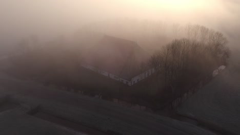 Traditional-dutch-farm-building-in-Friesland-countryside-during-low-morning-fog,-aerial