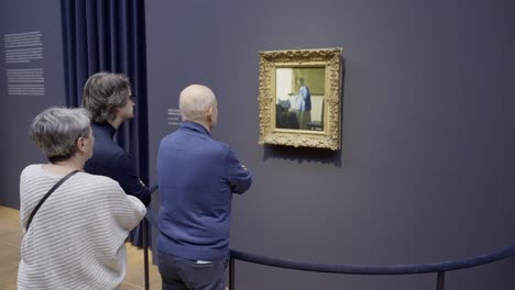 Begeisterte-Besucher-Bestaunen-Vermeers-„Die-Milchmagd“-In-Der-Vermeer-Ausstellung-Des-Rijksmuseums,-Anmutig-Gerahmt-Vor-Einem-Ruhigen-Blauen-Hintergrund