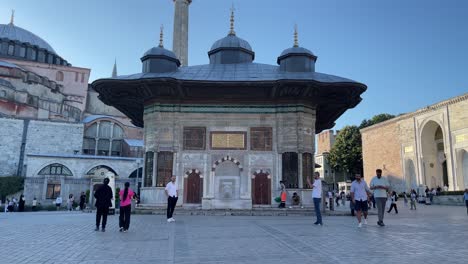 Frontal-view-of-the-Fountain-of-Ahmed-III-and-tourists-taking-photos-and-exploring-outside-the-imperial-gate-of-Topkapi-Palace-and-against-the-background-of-the-Hagia-Sophia-Mosque-in-Istanbul,-Turkey