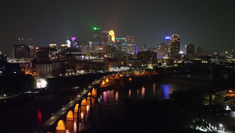 Blick-Auf-Die-Beleuchtete-Skyline-Der-Stadt-Minneapolis,-Nachtantenne-über-Dem-Mississippi-River