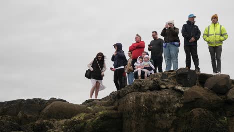 Spectators-watch-wearing-thick-jackets-standing-on-large-rocks,-grey-sky-behind
