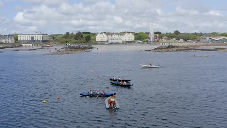 Drone-wide-angle-view-of-currach-boats-lined-up-at-start-line,-ladies-beach-galway-behind