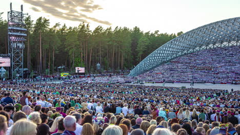 Timelapse-shot-of-huge-crowd-of-locals-gathering-to-watch-the-Latvian-Song-and-Dance-Festival-in-an-outdoor-stadium-in-Riga,-Latvia-with-sun-setting-in-the-background