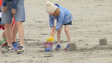 Young-child-wears-bright-blue-jacket-and-uses-shovel-to-fill-bucket-for-sand-castle