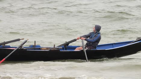 Solo-man-in-rain-jacket-smiles-as-he-rows-backwards-in-currach-boat
