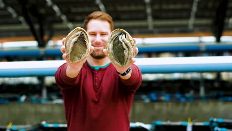 Smiling-caucasian-man-holds-up-two-big-South-African-abalones,-aquafarming