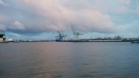 Hamburger-Hafen-Hafenkräne-Mit-Fliegenden-Möwen-Und-Vorbeifahrendem-Boot-Bei-Sonnenuntergang,-Blaue-Stunde,-Sanfte-Beleuchtung