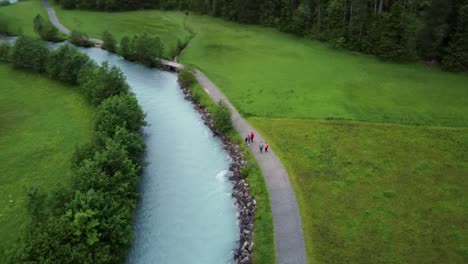 Tourist-People-taking-a-walk-by-a-glacial-river-bank-in-the-swiss-village-of-Kandersteg-amid-alpine-scenery-out-of-a-Fairy-tale-of-green-meadows,-forest-and-wooden-cabins