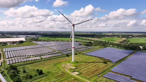 A-wind-turbine-standing-in-a-solar-field,-generating-renewable-energy---light-clouds,-blue-sky,-green-fields,-photovoltaic-power-plant