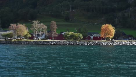 Blick-Von-Einem-Tiefblauen-Fjord-Auf-Eine-Kleine-Norwegische-Stadt-Am-Ufer-Mit-Den-Typischen-Roten-Hütten