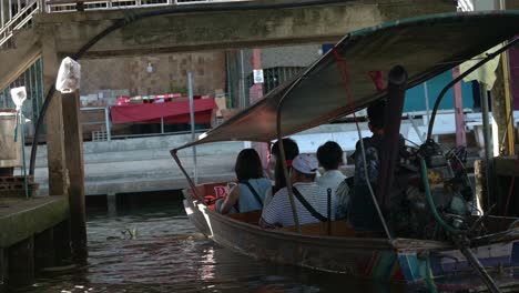 Wooden-boat-in-the-canal-of-Damoen-Saduak-Floating-Market-loaded-with-excited-tourists,-Thailand