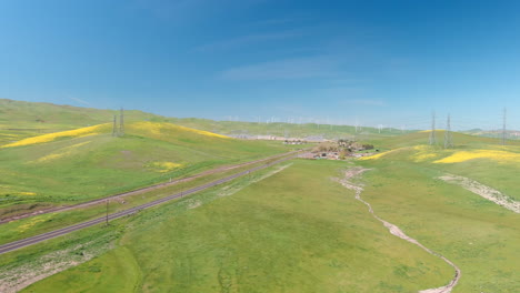 Large-powerlines-cross-a-highway-as-wind-turbines-spin-in-the-background-on-green-hills-dotted-with-yellow-wildflowers,-aerial
