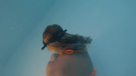 Cinematic-shot-of-a-woman-in-an-orange-bikini-and-vintage-diving-goggles-moving-gracefully-underwater-in-clear-blue-waters-shot-in-4K,-120-FPS,-Slomo