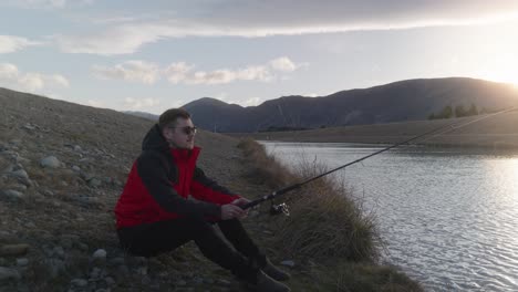 Young-man-with-red-jacket-fishing-sitting-down-at-calm-river-during-sunset