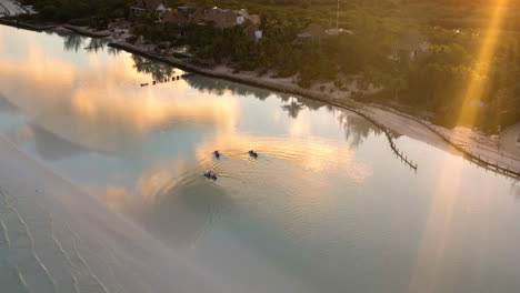 People-paddling-kayaks-along-beach-at-sunset,-clouds-reflecting-in-sea