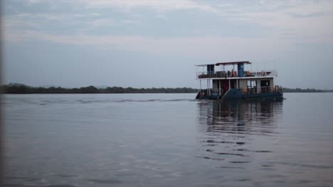Large-houseboat-ferry-slowly-drives-through-river-at-dusk,-slow-motion
