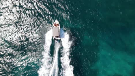 Aerial-top-down-rising,-small-boat-speeds-in-turquoise-blue-ocean-water,-tourist