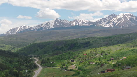 Vista-Aérea-De-Primer-Plano-A-La-Derecha-De-La-Hermosa-Cordillera-De-Colorado-Con-Picos-Nevados-En-Un-Día-Soleado-De-Cielo-Azul-En-El-Verano-Con-Campos-Verdes,-árboles-Y-Casas-De-Montaña
