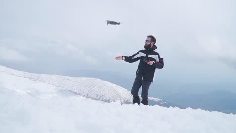Man-with-beard-launches-drone-from-hand-while-standing-in-snow-high-on-mountain