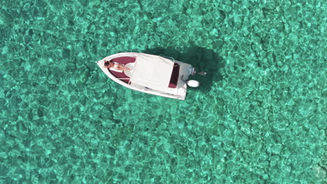 Top-Down-Aerial-View-of-Woman-in-Bikini-Sunbathing-on-Boat-Anchored-in-Crystal-Clear-Sea-Water