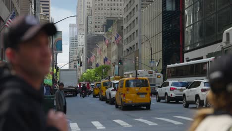 Gente-Esperando-Para-Cruzar-Una-Calle-Concurrida-De-La-Ciudad-De-Nueva-York-Con-Banderas-Americanas-Y-Tráfico-De-Automóviles.