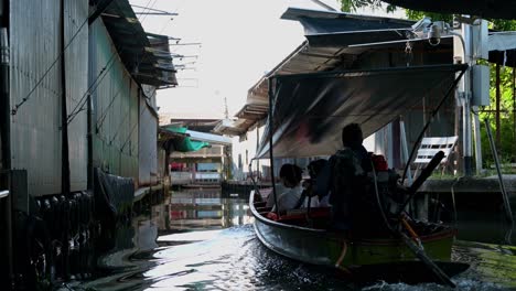 A-boatman-ferrying-a-group-of-tourists-in-a-wooden-boat-in-the-canals-of-Damnoen-Saduak-Floating-Market,-passing-by-the-closed-souvenir-shops-along-the-canal