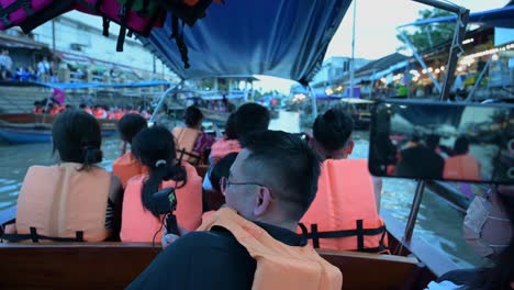 Groups-of-tourists-taking-a-boat-ride-in-Amphawa-Floating-Market-are-busy-with-taking-pictures-and-videos-while-sightseeing,-as-they-pass-by-restaurants-and-souvenir-shops-along-the-sides-of-the-canal