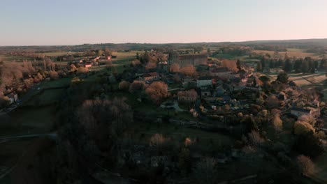 Aerial-view-with-elevation-of-a-small-village-and-its-church-in-the-middle-of-the-forest-at-sunrise,-Saint-Avit-Ségnieur,-Dordogne