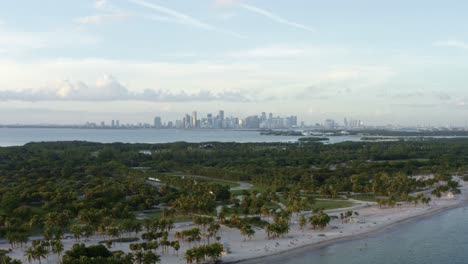 Aerial-drone-shot-of-the-beautiful-tropical-beach-surrounded-by-palm-trees-on-Crandon-Park-in-Key-Biscayne-with-the-skyline-of-Miami,-Florida-in-the-distance-on-a-warm-sunny-summer-evening