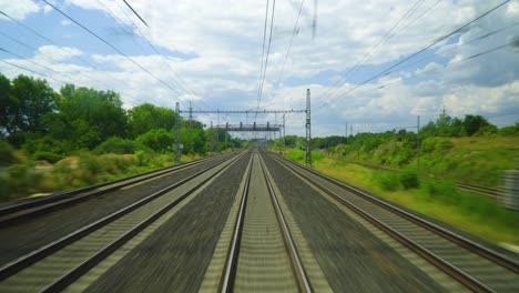 train-track-with-power-lines-and-trees-in-the-background-the-tracks-are-surrounded-by-lush-green-grass-and-there-is-an-overhead-power-line-running-along-the-length-of-the-track-white-clouds-scattered
