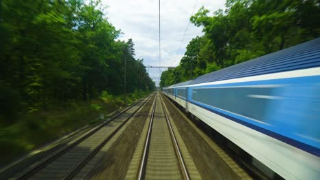 train-on-the-railway-tracks-surrounded-by-lush-green-trees-and-a-bright-blue-sky-train-is-traveling-opposite-the-track-with-power-lines-above-it-tall-trees-are-casting-shadows-onto-the-ground-below