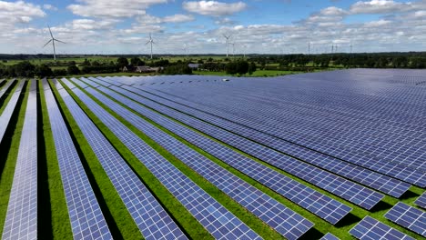 Rows-of-photovoltaic-solar-panels-in-a-solar-farm-generating-clean-and-eco-friendly-energy,-aerial-view,-drone-flight,-wind-turbines-in-the-background